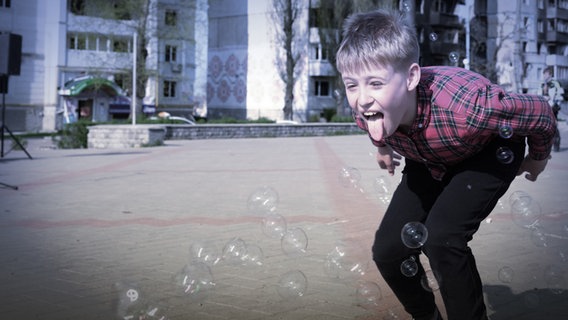 Ein Junge versucht mit seiner Zunge Seifenblasen einzufangen. Im Hintergrund sind Ostblock-Bauten zu sehen. © WDR/Mila Teshaieva 