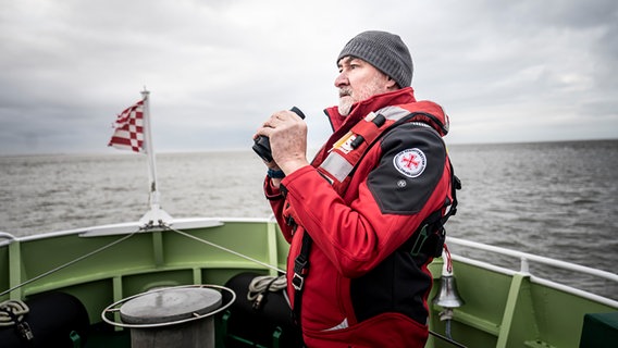 Ein Seenotretter steht an Deck und blickt aufs Meer. Er hält ein Fernglas in der Hand. © Kinescope/Achim Multhaupt 