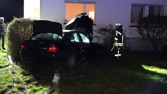 Ein Auto mit kaputter Front steht vor einem Haus, ein Feuerwehrmann steht neben dem Fahrzeug, ein Schlauch liegt auf dem Boden. © Nord-West-Media TV 