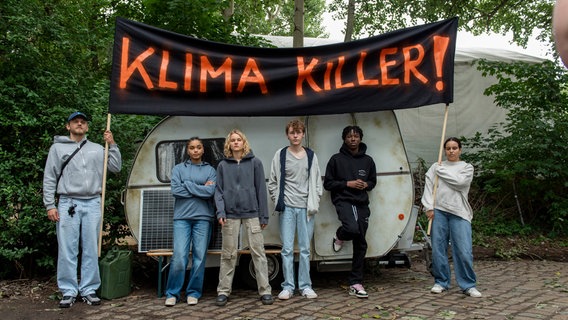 Sechs junge Aktivisten der Klimakiller-Killer stehen vor einem Wohnwagen und halten ein Transparent mit dem Text "Klima Killer" hoch. © ARD/Thorsten Jander 