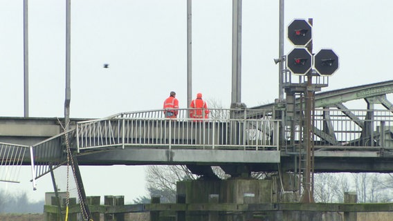 Zwei Personen in Signalarbeitskleidung stehen auf der beschädigten Eisenbahnbrücke über die Hunte.  © Screenshot 