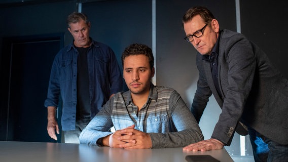 Der junge Jassim Abdaly (Mo Issa, M.) sitzt im Vernehmungsraum. Die beiden Ermittler Finn Kiesewetter (Sven Martinek, l.) und Lars Englen (Ingo Naujoks, r.) stehen hinter ihm. © ARD/Thorsten Jander 