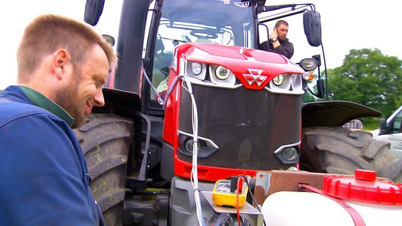 Ein Mann steht vor einem Traktor und schaut auf ein Gerät. Im Traktor steht ein Mann in der geöffneten Tür und sieht zu. © NDR 