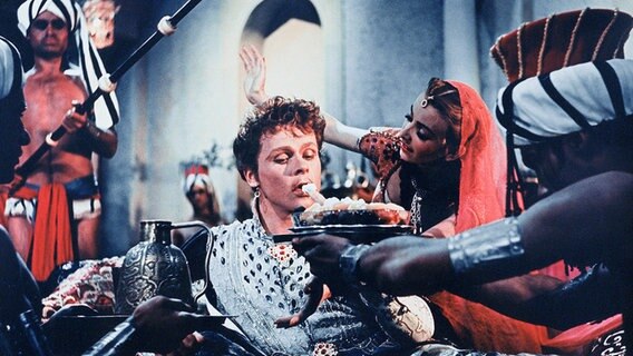 Ein junger Mann in einem Prinzengewand wird mit Essen und Trinken versorgt. Eine junge Frau tanzt im Hintergrund. © MDR 