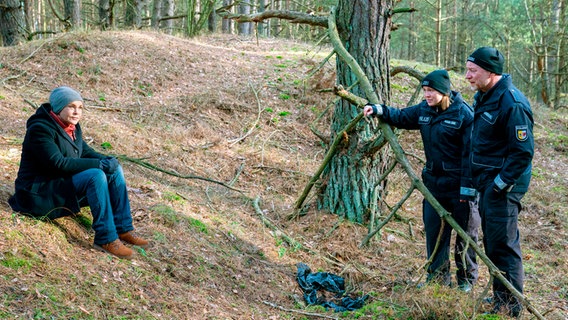 Eine Frau sitzt auf dem Boden in einem Wald. Ihr gegenüber stehen eine Polizistin und ein Polizist hinter einem herunterhängenden Ast. © NDR/ARD Degeto/Oliver Feist Foto: Oliver Feist