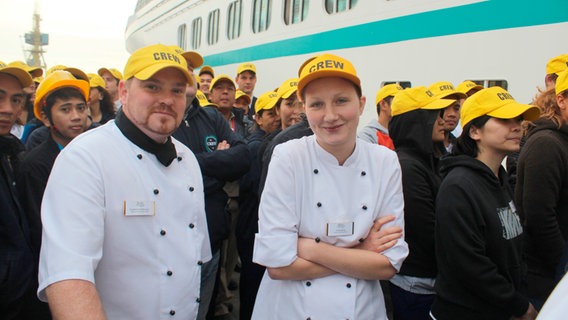 Eine Gruppe Menschen, die gelbe Kappen mit der Aufschrift "Crew" tragen, stehen vor einem Kreuzfahrtschiff. © BR/Bewegte Zeiten GmbH/Gerrit Manne Foto: Gerrit Manne