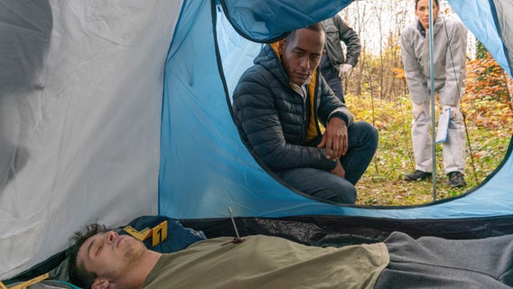 In einem Zelt liegt ein toter junger Mann. Mehrere Personen befinden sich vor dem Zelt und schauen hinein. © ARD/Lucky Bird Pictures/Angela M. Schlabitz Foto: Angela M. Schlabitz