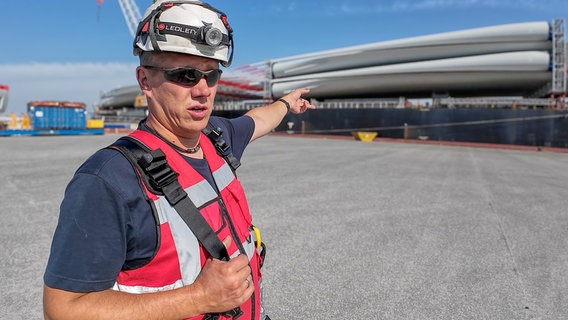 Hafenarbeiter Malte Hoffmann zeigt auf einen Schwertransporter mit Bauteilen für Windräder. © NDR/FILMBLICK Hannover/Martin Göbel 