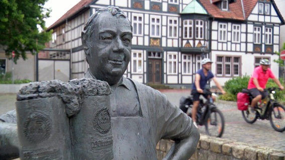 Zwei Männer fahren mit dem Rad eine Straße entlang, im Vordergrund ist die Skulptur eines Bierbrauers zu sehen. © NDR 