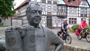 Zwei Männer fahren mit dem Rad eine Straße entlang, im Vordergrund ist die Skulptur eines Bierbrauers zu sehen. © NDR 