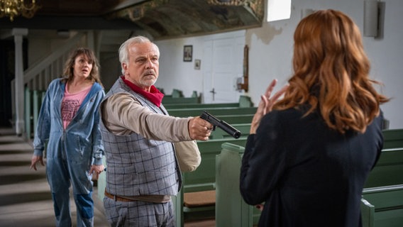 Szenenbild aus der NDR Serie "Reisen mit Muddi": Ein Mann bedroht eine Frau mit einer Waffe, eine andere Frau verfolgt die Szene. © NDR/Boris Laewen Foto: Boris Laewen