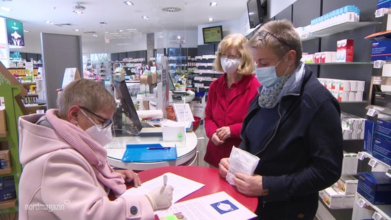 Three women are standing in the pharmacy.  Everyone is wearing masks.  