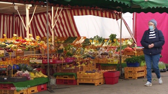 View of the market stalls of a weekly market.  A woman walks through the picture from the right, wearing mouth and nose protection.  