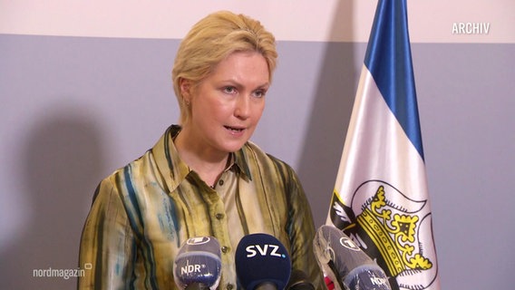 Prime Minister Schwesig at a press conference, a flag behind her, microphones on a table in front of her.  