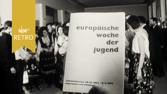 Programmheft "europäische woche der jugend" wird vor eine Gruppe von Jugendlichen in verschiedenen Trachten gehalten (1964)  
