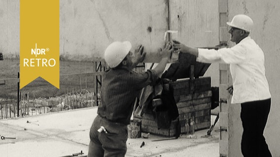 Bauarbeiter dirigieren eine Stahlbeton-Fertigwand auf der Bausstelle (1964)  