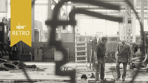 Bauarbeiter in einer Messehalle in Hannover, hinter einem Logo 1964  