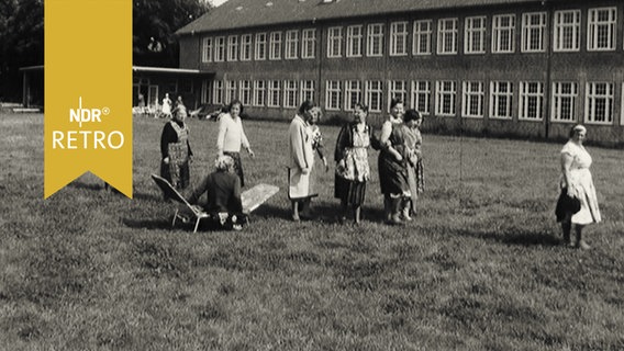 Frauen vor einem zweistöckigen Wohnheim, offenbar von der Gartenarbeit zurückkehrend (1964)  