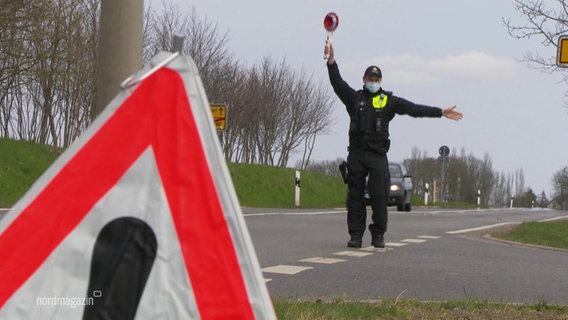 At the edge of a road a policeman holds up a stop-trowel, in the foreground there is a warning triangle.  