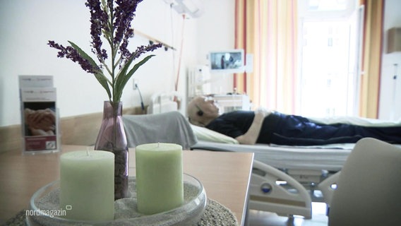 A patient room in the hospital, in the background an old man is lying on the bed, in the foreground there are two candles on a table.  