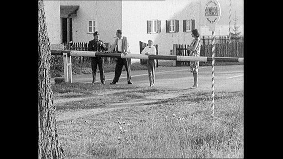 Vier Personen stehen an einer Grenzschranke.  