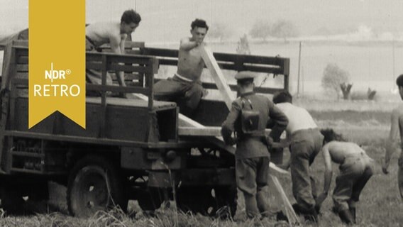 Mitglieder der DDR-Volkspolizei, teils mit nacktem Oberkörper, laden Balken zur Grenzbefestigung von einem Armeelaster (1963)  