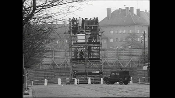 Menschen stehen auf einer Aussichtsplattform an der Bernauer Straße und schauen über die Mauer.  