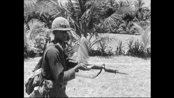 Ein Soldat mit Gewehr läuft auf einer Wiese mit Palmen.  