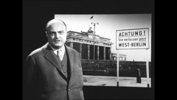 Der Publizist Sebastian haffner steht vor einem Foto des Brandenburger Tors, davor das Schild "Achtung, sie verlassen jetzt Westberlin"  