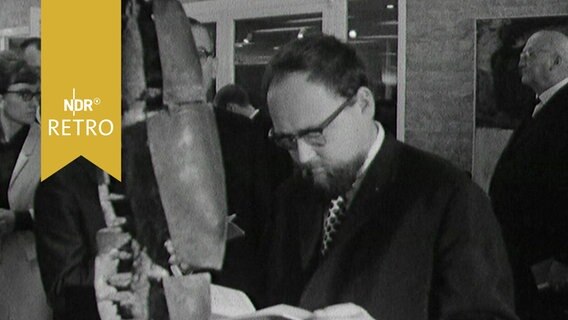 Bärtiger Mann steht vor einer Schrottskulptur aus Metall und liest in einem Begleitheft - in einer gut besuchten Ausstellung in Wolfsburg 1963  