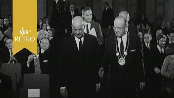 Göttinger Universitätsdirektor Arnold Scheibe und Professor Clark Kerr ziehen zu einer Festveranstaltung in einen Saal ein (1963)  