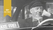 Älterer Taxifahrer wartet am Steuer seines Taxis auf Kundschaft, im Fenster ein Schild "Taxe frei" (1958)  