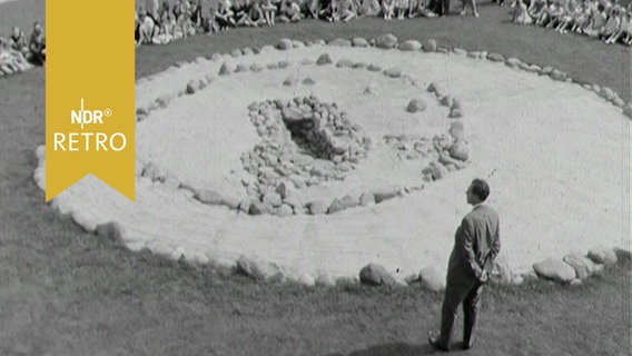 Besucher sitzen im Kreis um eine runde Fläche, die ein Steinzeitgrab darstellt (1963)  