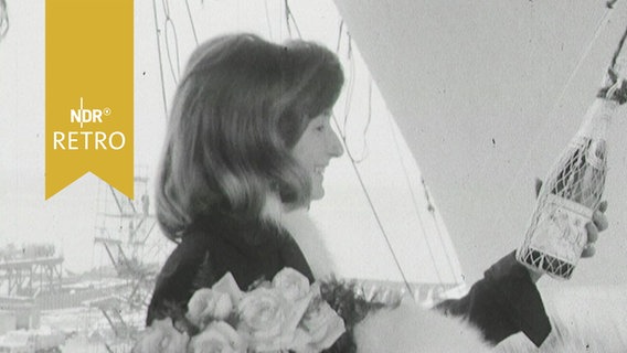 Junge Frau hält vor einer Schiffstaufe die Sektflasche in der Hand und lächelt (1964)  