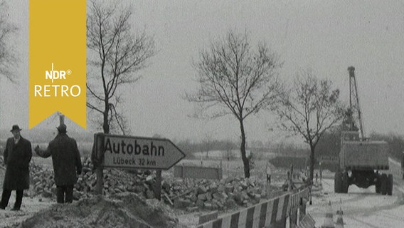 Straßenbaustelle auf dem Land: Neben einem Wegweiser zur "Autobahn" steht ein Bagger (1964)  