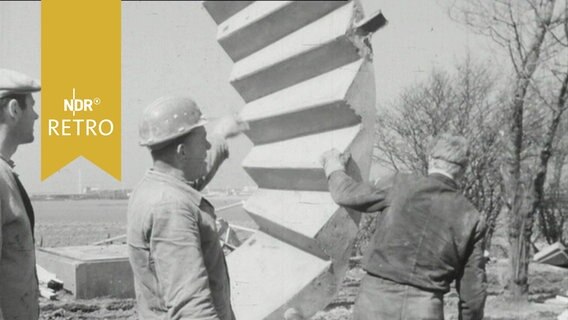 Arbeiter dirigieren ein an einem Kran hängendes Betontreppenelement (1964)  