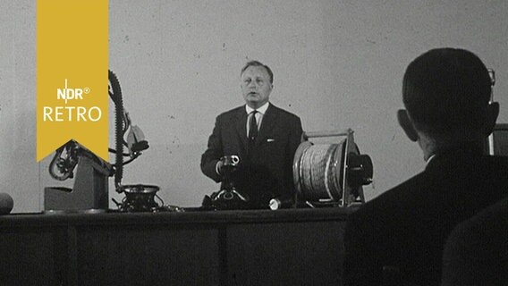 Ein Vortragender auf der Jahrestagung des Brandschutz-Vereins 1964 zeigt Schutzausrüstung und eine Seilwinde am Podiumstisch  
