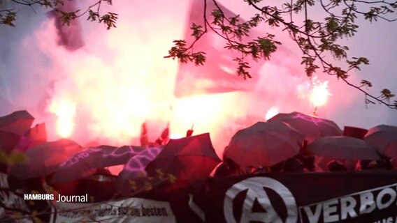 Hinter einem Banner laufen Menschen, die sich hinter schwarzen Regenschirmen verschanzen, im Hintergrund roter Nebel und Bengalos. © Screenshot 