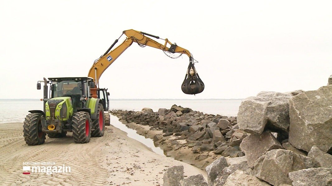Ein Bagger hebt einen schweren Stein um ihn auf einer Steinpallisade an einem Strand abzusetzen.