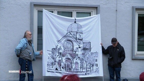 Zwei Männer halten ein Transparent, auf dem eine Synagoge gezeichnet ist. © Screenshot 
