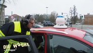 Ein Mann mit Kopfhörern und Signalweste neben einem Auto, das eine Sirene auf dem Dach installiert hat. © Screenshot 
