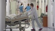 Eine Frau in weißer Arbeitskleidung schiebt auf einem Gang eines Krankenhauses ein Krankenbett in einen Seitengang. © Screenshot 