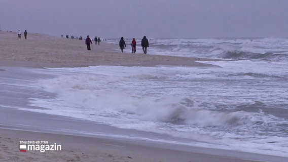 Die Urlauber auf Sylt lassen sich auch von Wind und Wetter nicht abschrecken und gehen am Strand spazieren. © Screenshot 