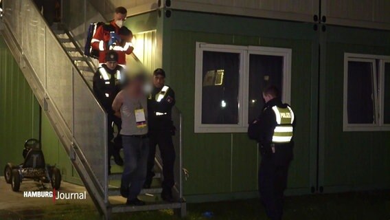 Einsatzkräfte der Polizei führen einen unkenntlich gemachten Mann bei Nacht eine Außentreppe einer Containersiedlung in einer Unterkunft für Geflüchtete hinab. © Screenshot 