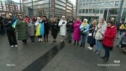 Eine Gruppe von Menschen steht bei einer Friedensdemo singend im Halbkreis mit einer Ukrainefahne. © Screenshot 