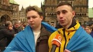 Zwei junge Erwachsene tragen eine Ukraine-Fahne um den Körper und sprechen in ein Mikrofon. © Screenshot 