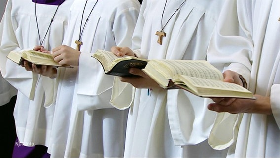 Mehrere Personen in Messdienertracht lesen in Bibeln und tragen Ketten mit Kreuzen um den Hals. © Screenshot 
