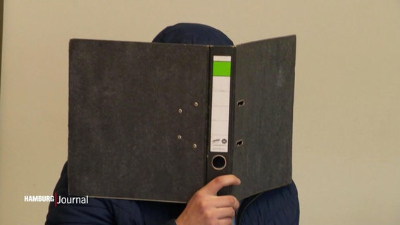 Ein Mann schützt sein Gesicht im Gerichtssaal mit einem Aktenordner vor den Kameras. © Screenshot 