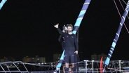 Segler Boris Hermann steht mit hochgehaltenem "Shaka"-Handzeichen auf dem Deck des in den Hafen einlaufenden Segelboots seiner Crew. © Screenshot 