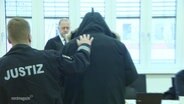 Ein Justizbeamter führt eine Person mit schwarzer Kapuzenjacke in einen Gerichtssaal. © Screenshot 